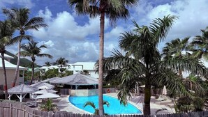 En swimmingpool omgiven av palmer och ett trästaket, med strandparasoller och en pergola i bakgrunden.