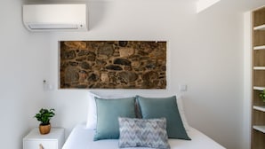 Ein Schlafzimmer mit einem Kunstwerk an der Steinwand, einem Bett mit zwei Kissen, einer Zimmerpflanze und einem weißen Nachttisch mit Schubladen.