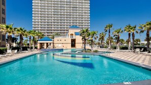 Ein Hotelpoolbereich mit einem großen Gebäude im Hintergrund, Palmen und Liegestühlen.