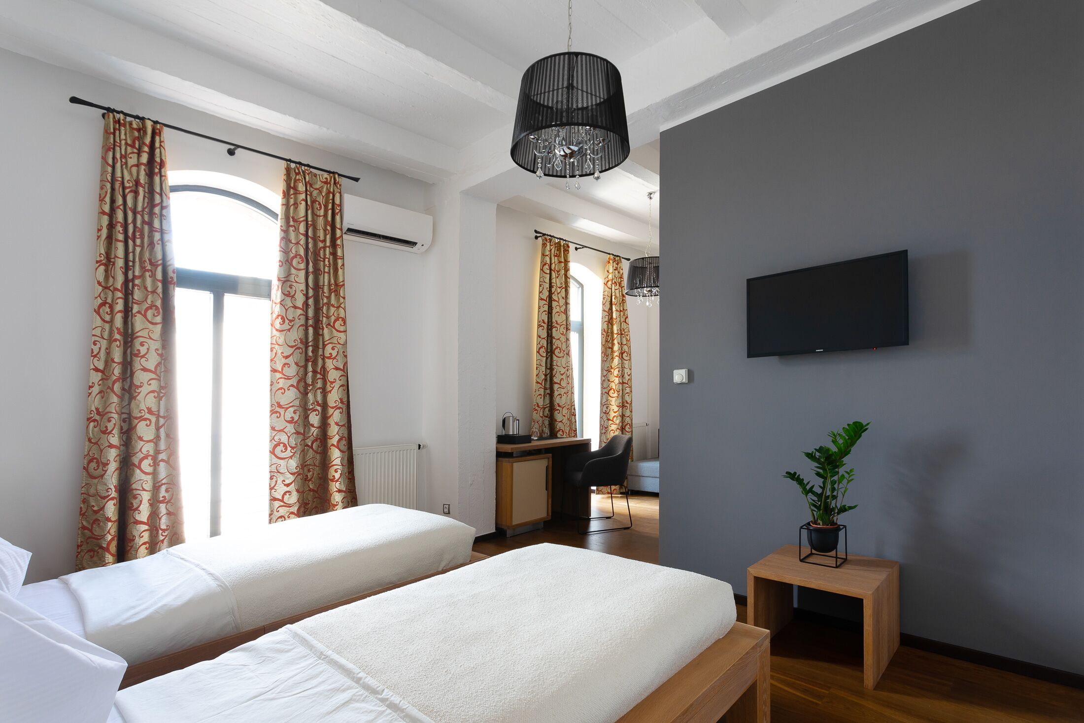 A modern hotel room with two beds, a desk, a chair, a television, and a potted plant.