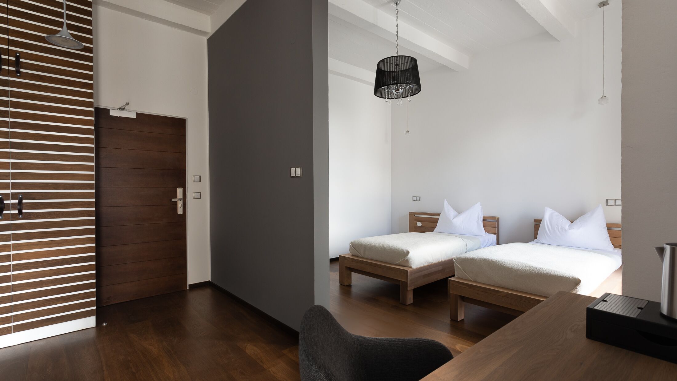 A modern hotel room with two beds, a wooden floor, a dark grey accent wall, and a hanging lamp.