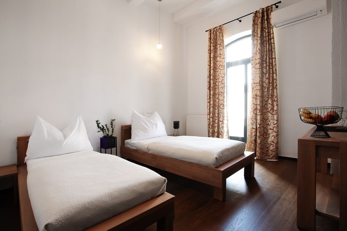 Um quarto com duas camas de solteiro, uma mesinha de cabeceira com luminária, uma pequena planta e uma janela com cortinas.