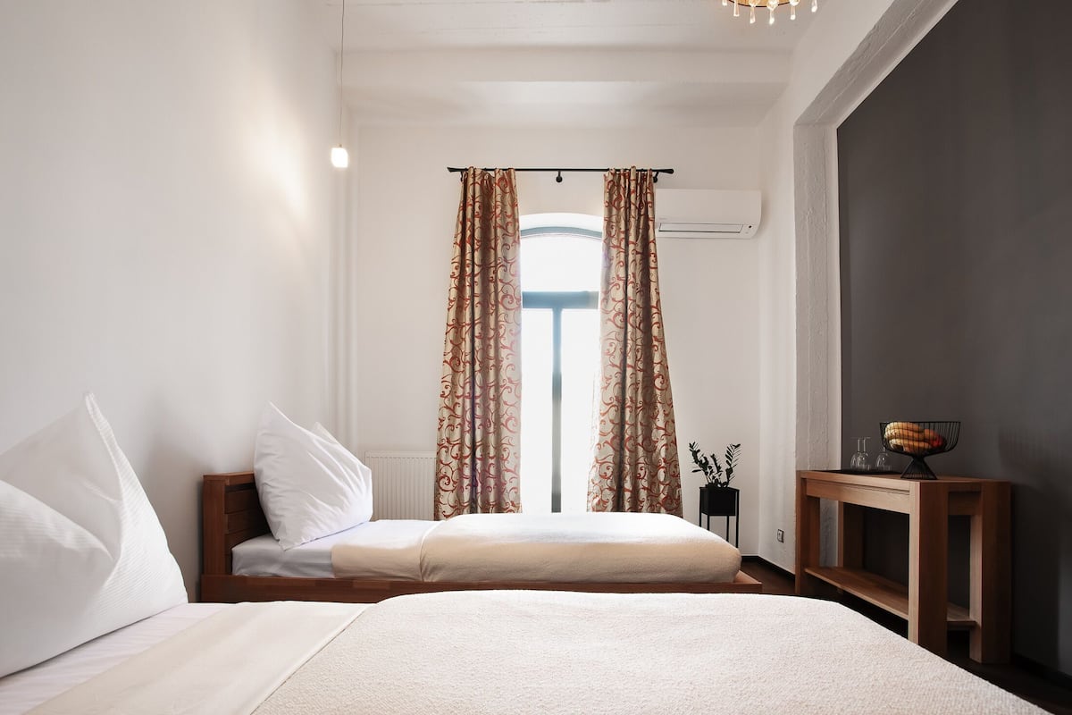 Quarto com duas camas, uma janela com cortinas, uma mesinha de cabeceira com uma tigela e um ar condicionado fixado na parede.