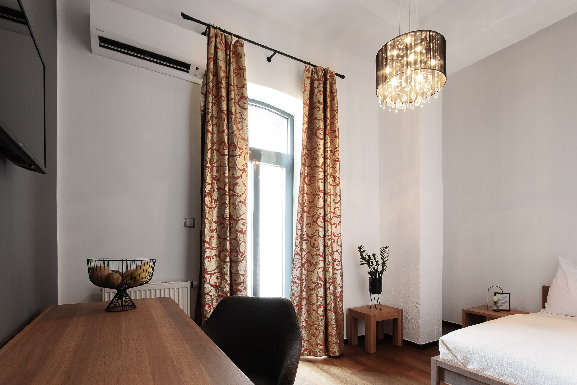 Una habitación de hotel moderna con una mesa de comedor de madera, una silla negra, una cama con ropa blanca, una mesita de noche pequeña y una lámpara de noche, una lámpara de araña colgante y una ventana con cortinas estampadas.