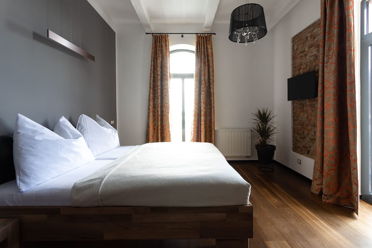 Quarto moderno com cama de madeira, roupa de cama branca, televisão montada e uma janela grande com cortinas.