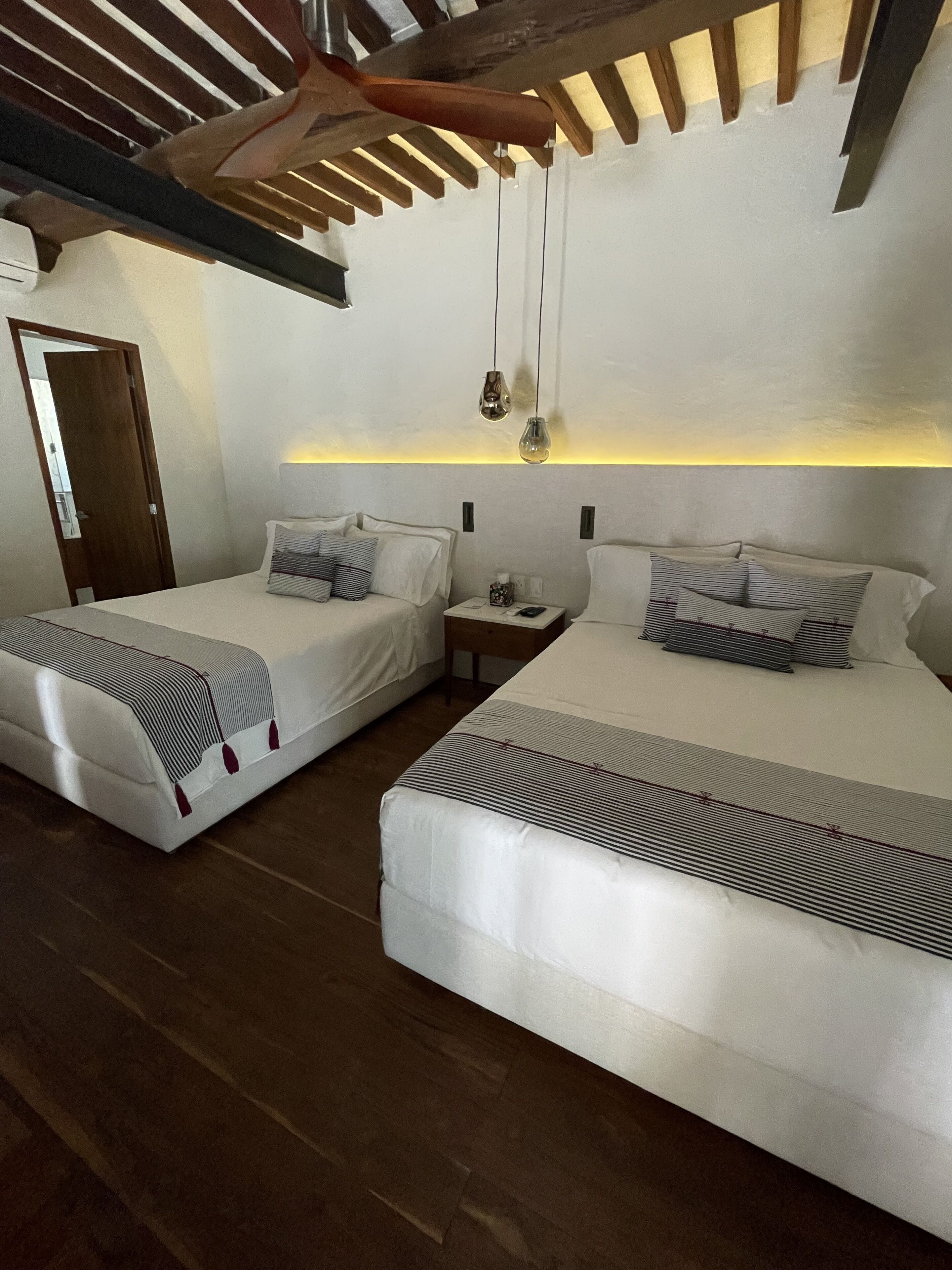 Two single beds with striped bedding, a bedside table with a lamp, and a wooden ceiling structure.