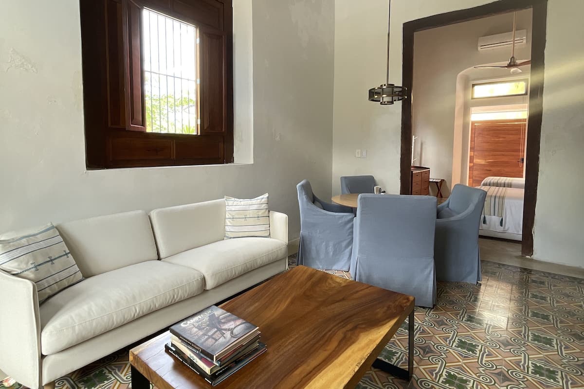 Una sala de estar con un sofá, una mesa de centro, baldosas de suelo estampadas, un ventanal con contraventanas de madera y un ventilador de techo.