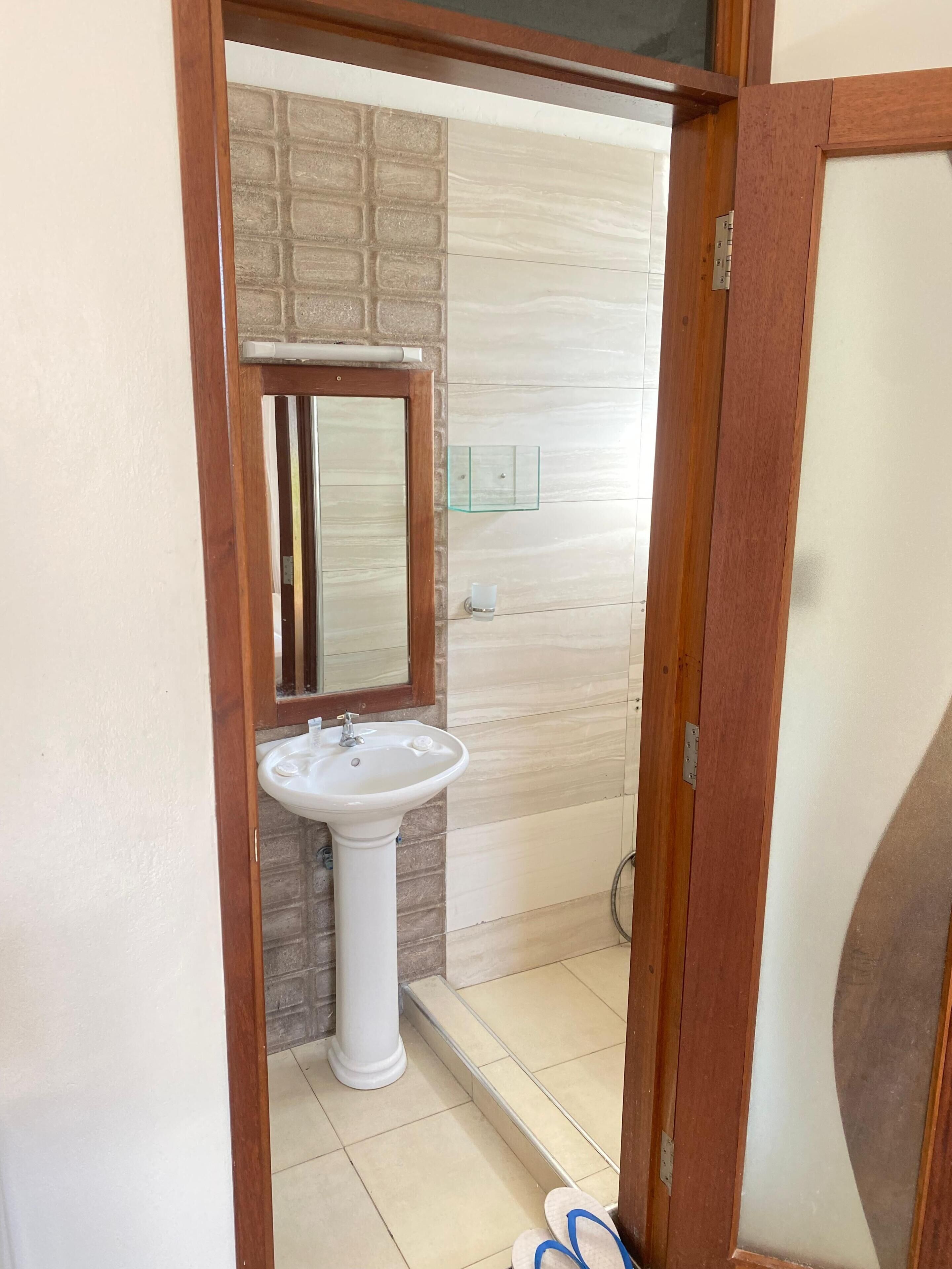 Casa de banho com lavatório pedestal branco, espelho e estante de vidro.