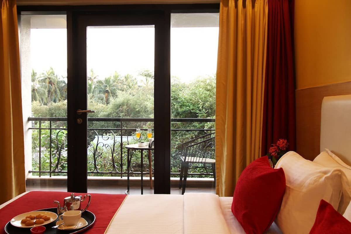 A hotel room with a balcony, a bed with red pillows, a tray with food and a teapot, and a view of greenery and a table with drinks.