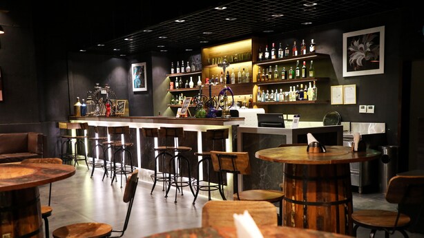 A bar area with wooden tables and chairs, a counter with a menu, and shelves with bottles. - Rosetum Anjuna (Anjuna)