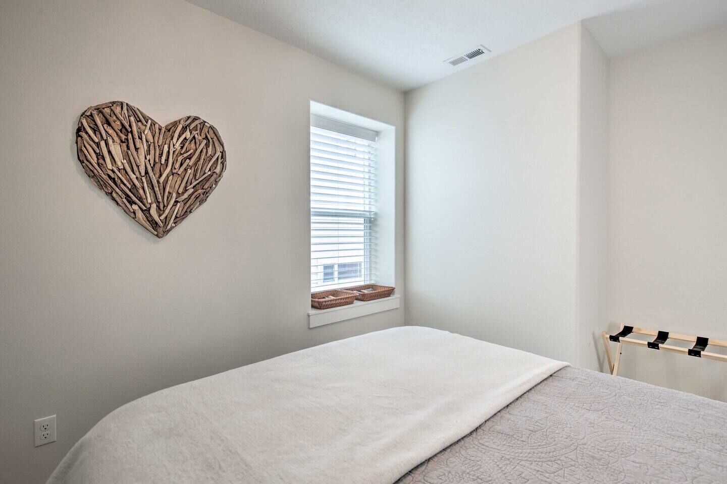 A bedroom with a bed, a heart-shaped wall decoration, a window with blinds, and a drying rack.