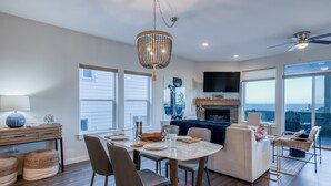 A spacious living room with a dining area, a fireplace, and large windows offering a view. - Ocean Views + Hot Tub | Walk Harbor | Crab Cooker | Sleeps 6 (Depoe Bay)