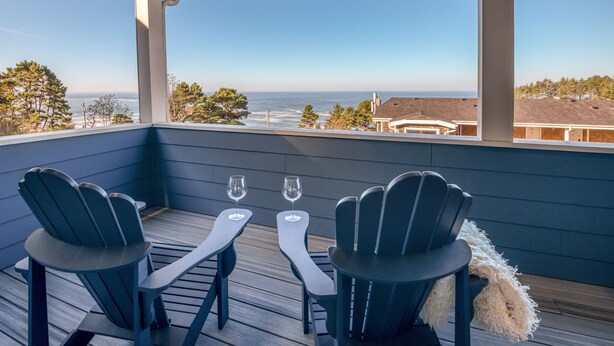 A covered deck with two Adirondack chairs and two wine glasses on a wooden floor, overlooking a beach and ocean. - Ocean Views + Hot Tub | Walk Harbor | Crab Cooker | Sleeps 6 (Depoe Bay)