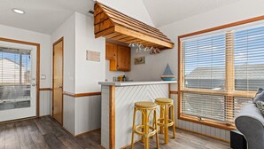 A compact kitchen with a wooden bar, two stools, and a view of the beach through large windows. - Totally Beach'n 4 Bedroom Home in Kill Devil Hills (Kill Devil Hills)