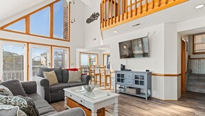 A spacious living room with a high ceiling, large windows, a wooden staircase, and a television mounted on the wall. - Totally Beach'n 4 Bedroom Home in Kill Devil Hills (Kill Devil Hills)