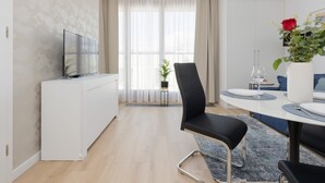 Uma sala de estar moderna e bem iluminada, com uma televisão de tela plana, um espaço de jantar com mesa branca e cadeiras pretas, e uma janela grande com cortinas translúcidas.
