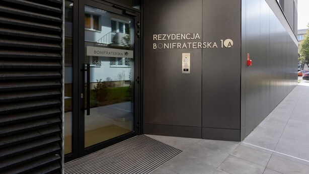 Uma fachada moderna de edifício com porta de vidro, portão de metal e uma placa com a inscrição "REZYDENCJA BONIFRATERSKA 1A".