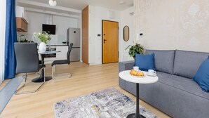 A modern living room with a grey sofa, a small round table with a plate of food, and a dining area with a black chair and a vase of flowers.