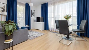A modern living room with a grey sofa, two black chairs, a small table, and a TV on a white cabinet. Blue curtains and a large window provide natural light.