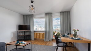 A modern living room with a TV, dining table, and chairs, large windows with curtains, and a city view.
