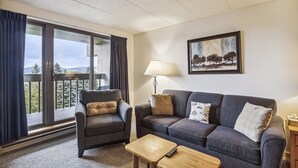 A living room with a sofa, armchair, and coffee table, a painting on the wall, and a balcony with curtains. - Pinnacle A11  Condo | Pool, Hot Tub | Golf Nearby (Killington)