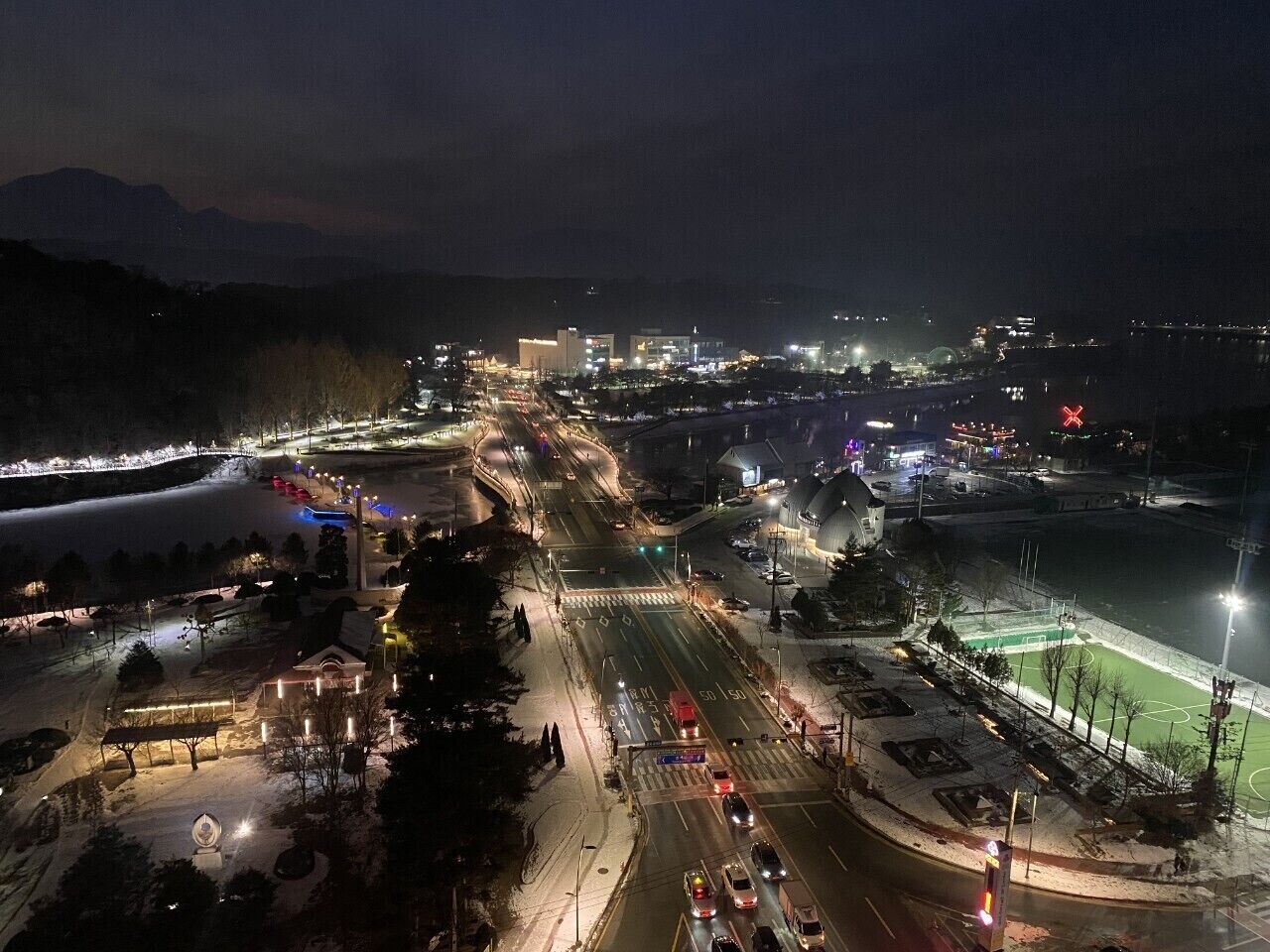 Une vue aérienne nocturne d’une ville, avec des rues illuminées, un terrain de sport et des bâtiments en arrière-plan.