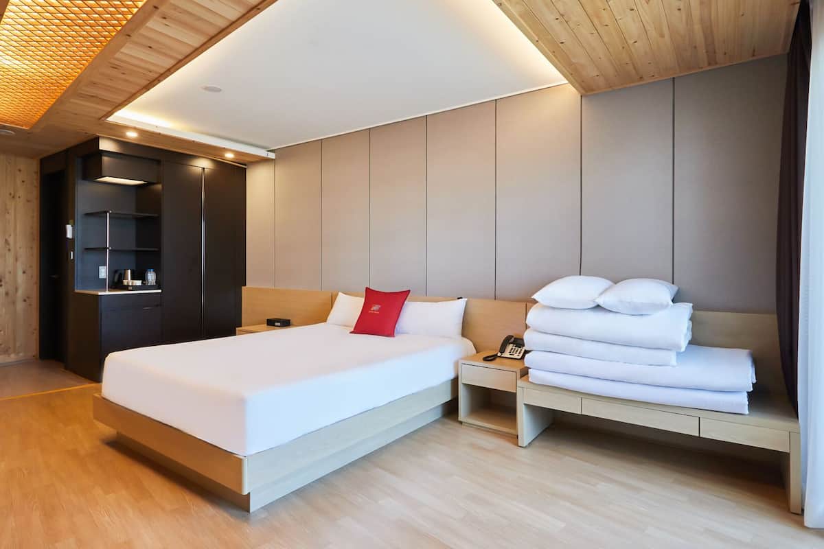 A modern hotel room with a large bed, a bedside table with a phone, a red pillow, and a stack of white towels.