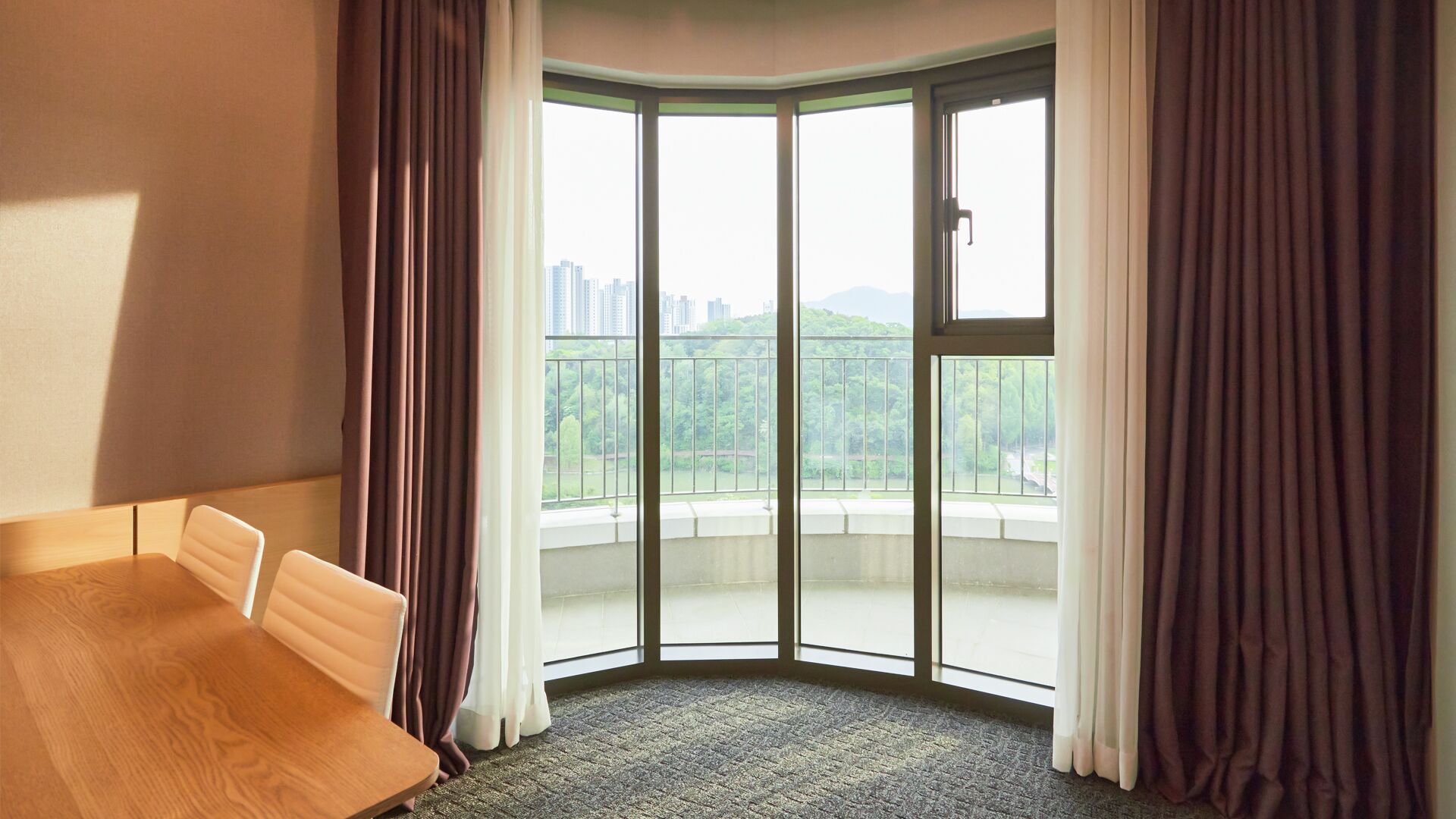 A hotel room with a corner bed, a view of a cityscape and greenery through a large glass door, and a balcony with a railing.