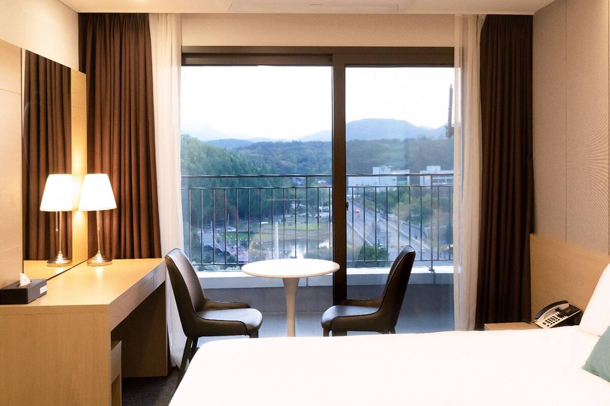 A hotel room with a bed, a desk with a lamp, two chairs, a small table, and a balcony with a view of a city and mountains.