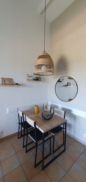 Uma área de refeições com mesa de madeira, cadeiras pretas, luminária pendente trançada, um vaso amarelo e um espelho redondo na parede.