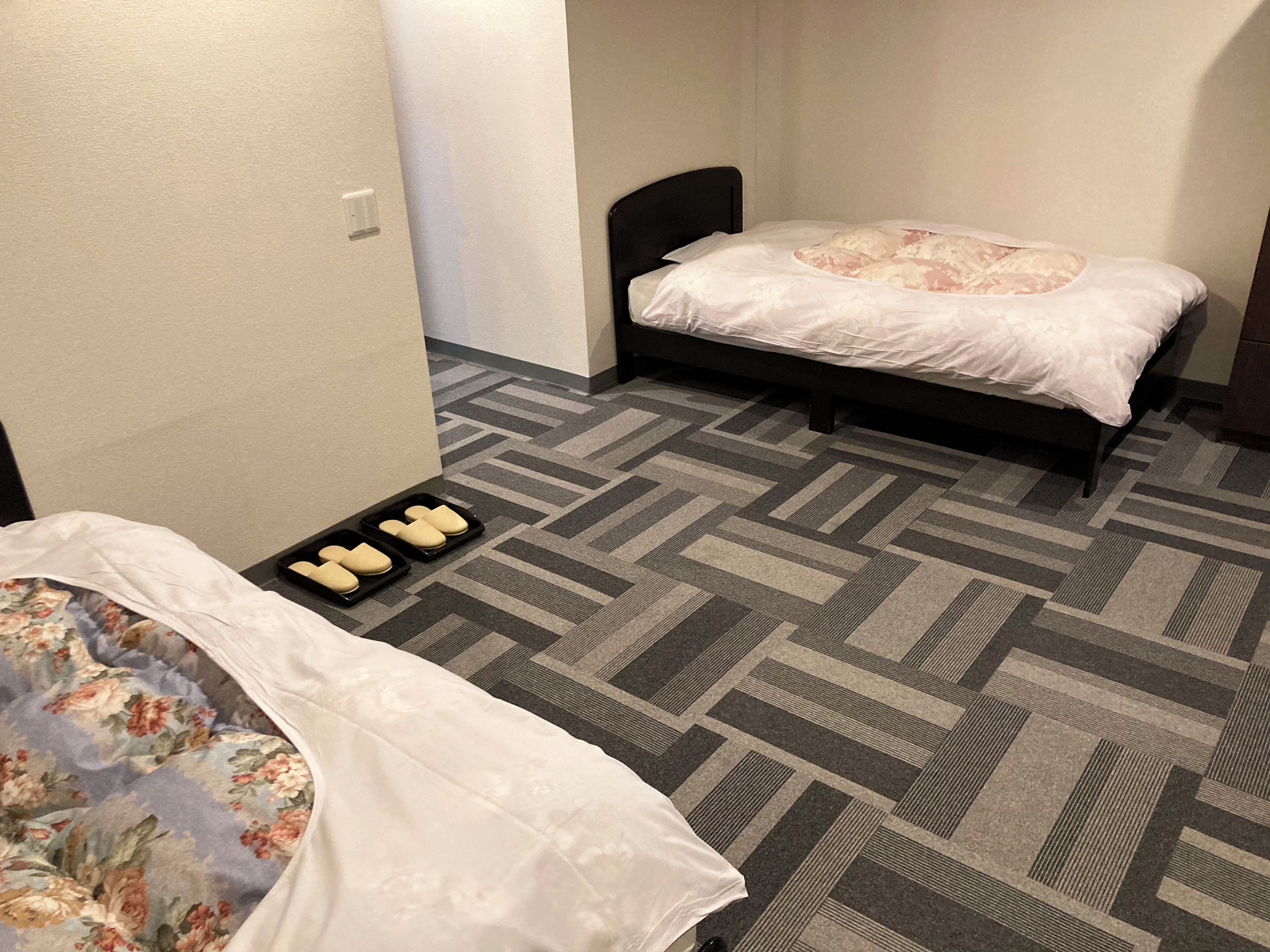 A hotel room with two beds, a patterned carpet, and a tray with slippers.