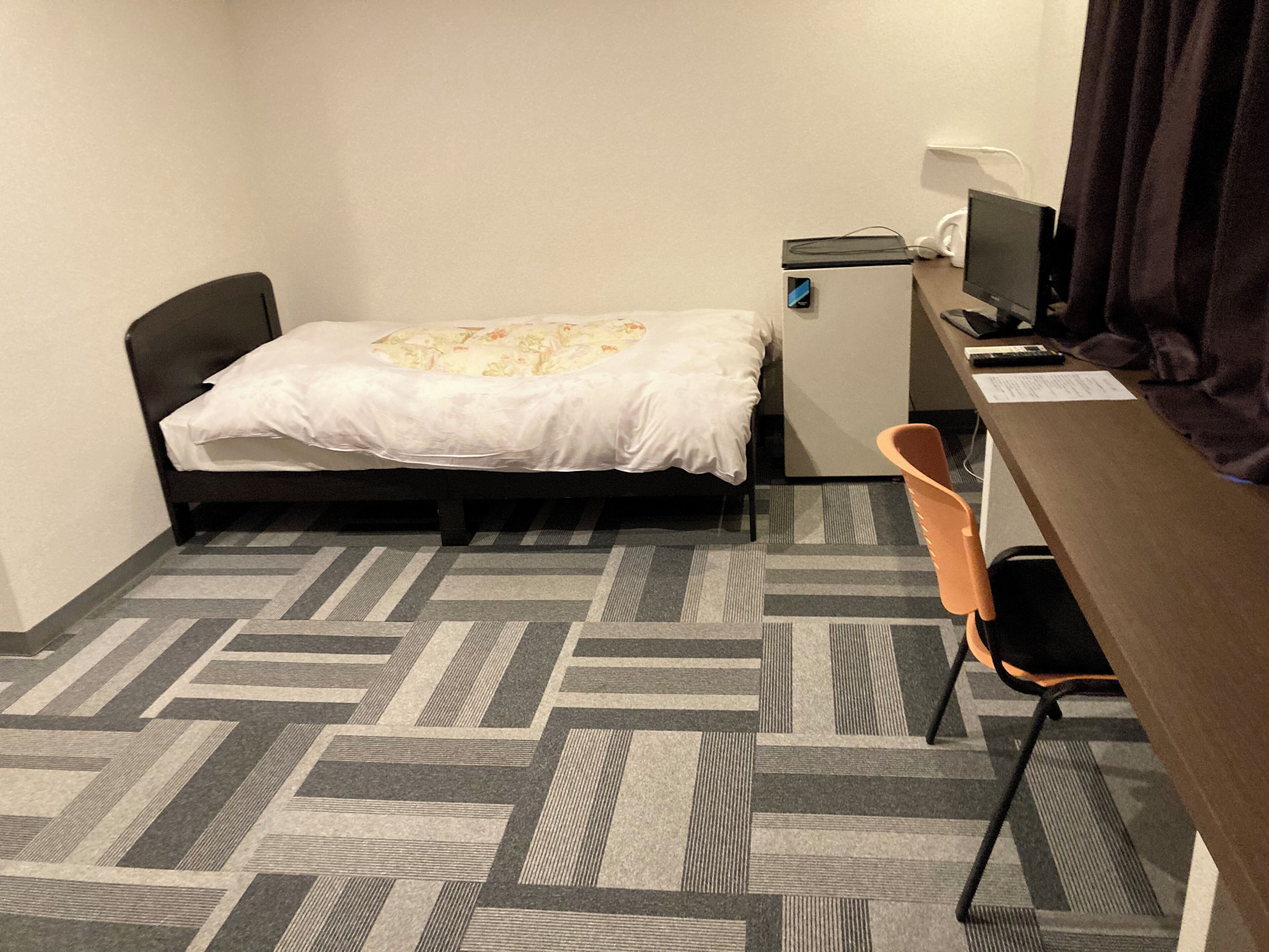 A small hotel room with a bed, a desk, a chair, a small refrigerator, and a television.