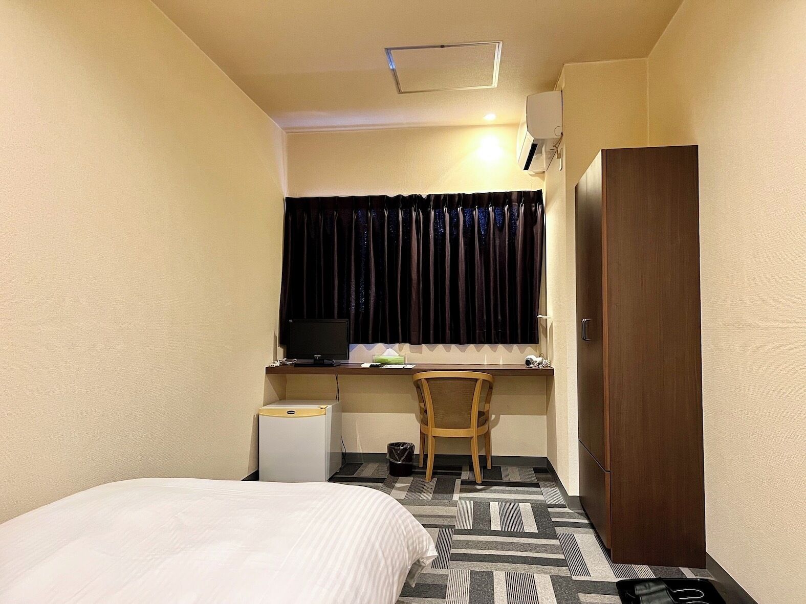 A small hotel room with a bed, a desk with a computer, a chair, a wardrobe, and a window with curtains.