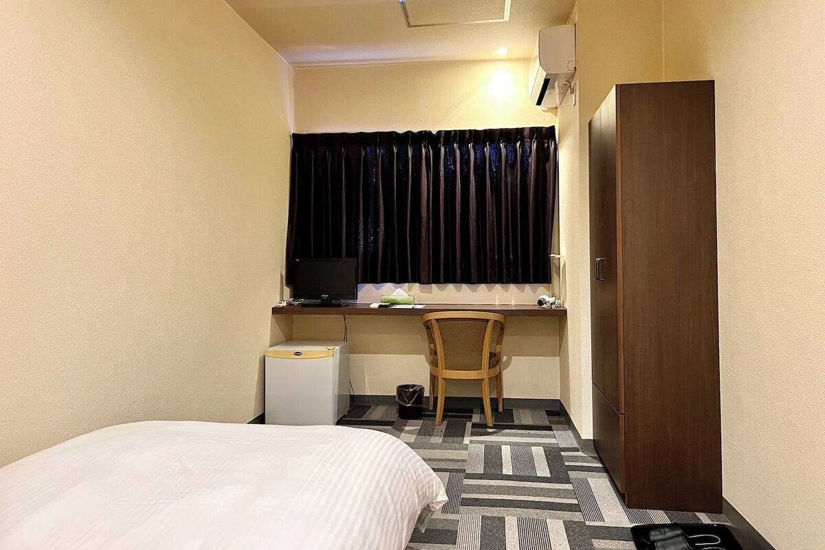 Een kleine hotelkamer met een bed, een bureau met een computer, een stoel, een kledingkast en een raam met gordijnen.