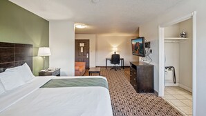 A hotel room with a bed, a desk, a chair, a television, and a coat rack. - Coratel Plus Luxury Suite 1 King bed with Sofa Non-Smoking Oversized (Wichita)