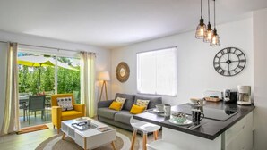 Ett modernt vardagsrum med en soffa, ett soffbord och stolar. Det finns en matplats med ett bord och stolar. Ett köksområde med en köksbänk och vitvaror syns. Ett stort fönster och en dörr som leder ut till ett utomhusområde kan också ses.
