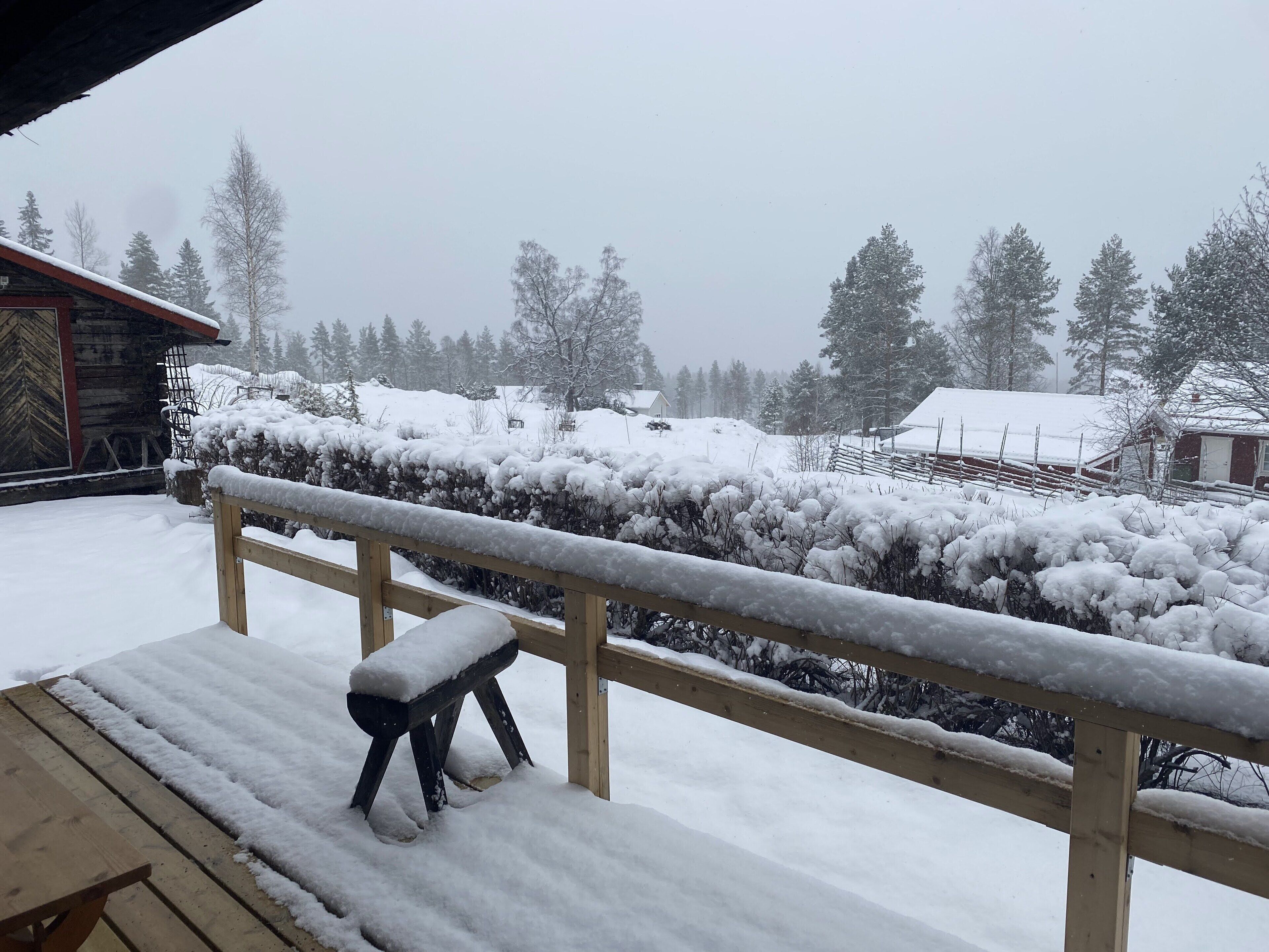 A wooden deck with a bench covered in snow, a railing, and a view of a snowy landscape with trees and buildings in the background.