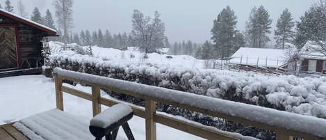 A wooden deck with a bench covered in snow, a railing, and a view of a snowy landscape with trees and buildings in the background.