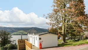 Un petit bâtiment blanc avec un escalier en bois et un extincteur rouge, situé près d’une étendue d’eau et d’un arbre aux feuilles d’automne.