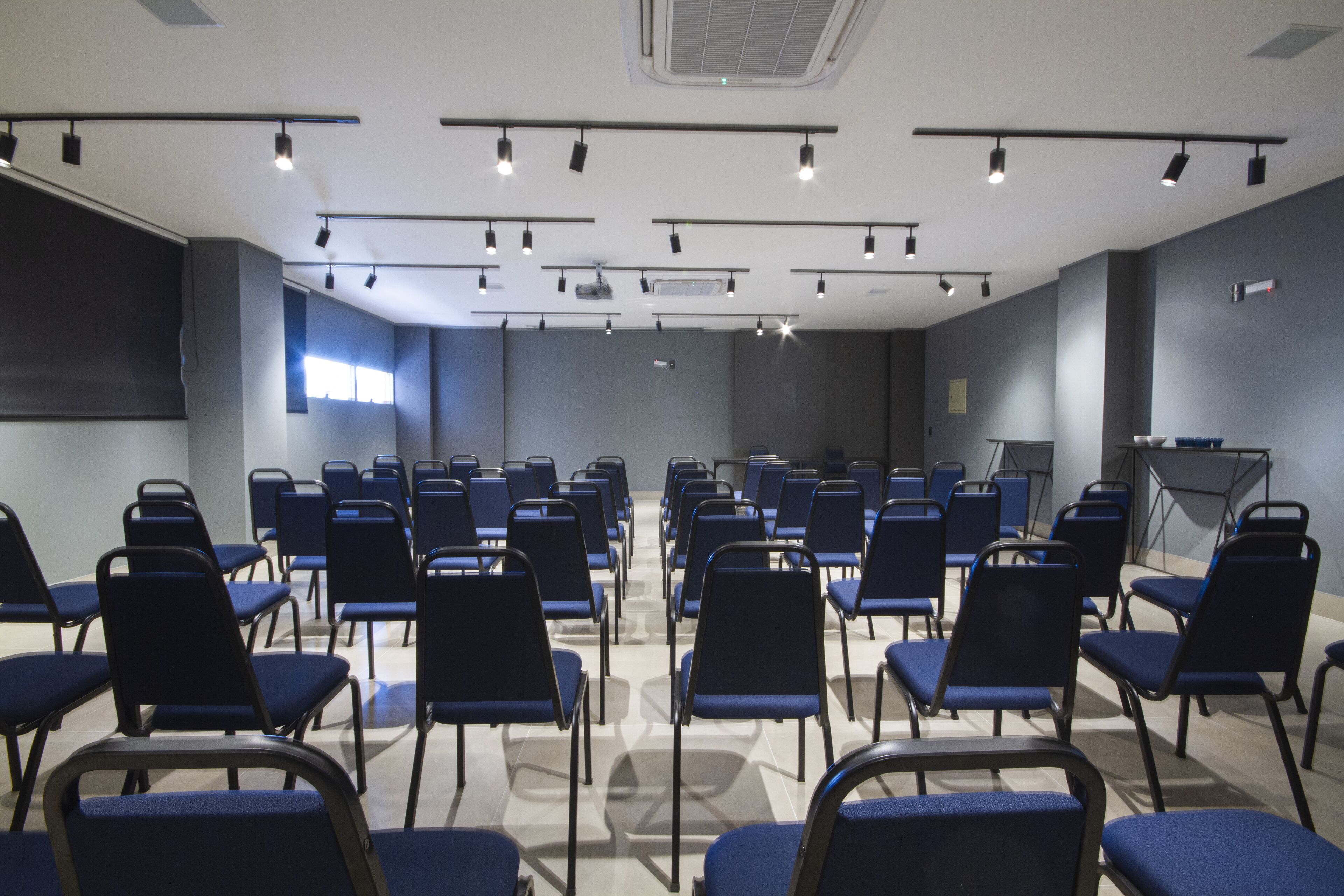 Un salón de conferencias con hileras de sillas azules, una pantalla de proyector y luces en el techo.