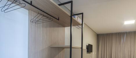 Una habitación de hotel compacta con una cama, un escritorio y una silla. Se observa un armario con ropa colgada y un televisor montado en la pared.