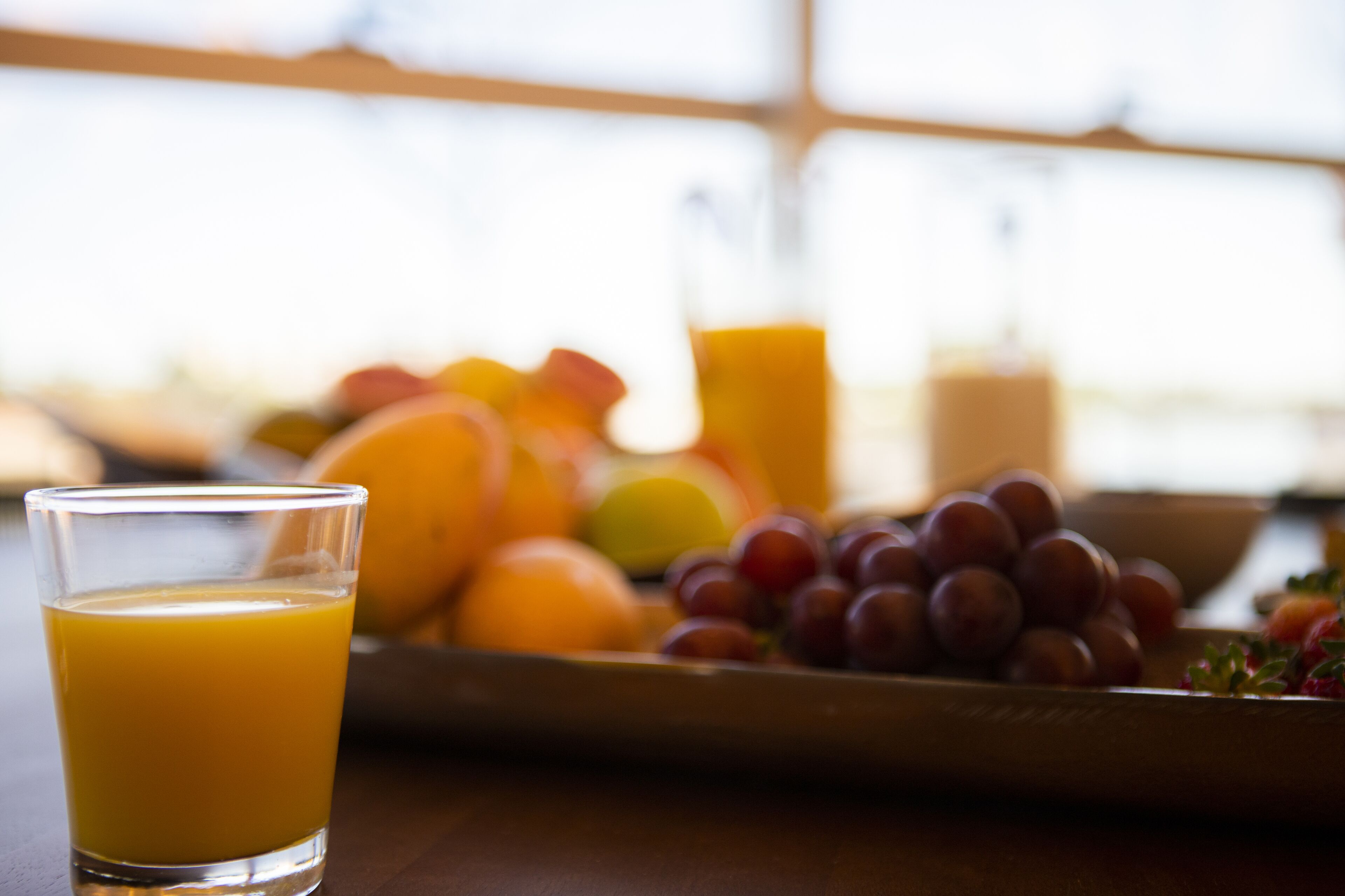 Un vaso de jugo de naranja sobre una mesa, con un bol de frutas al fondo.