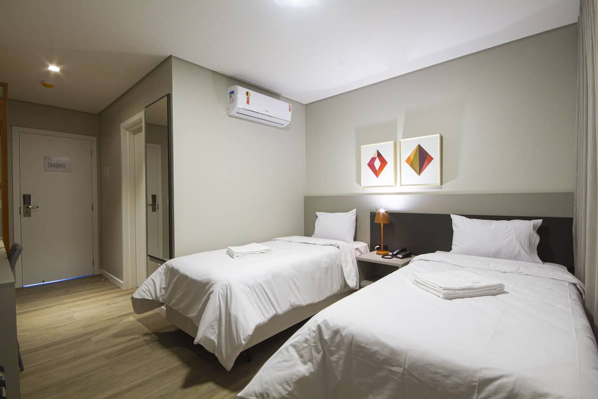 Una habitación de hotel con dos camas, una mesita de noche con lámpara, un equipo de aire acondicionado y una puerta con un letrero.