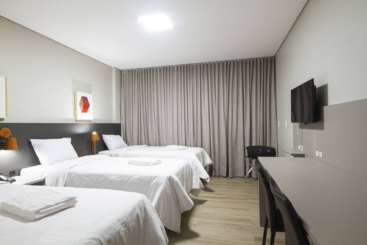 Habitación de hotel con tres camas, un escritorio, un televisor y un ventanal grande con cortinas.