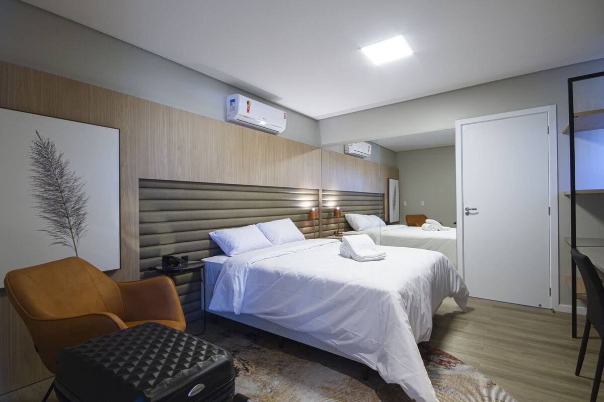 Habitación de hotel con dos camas, cabecero de madera, aire acondicionado de pared, lámpara de noche, una silla y una maleta.