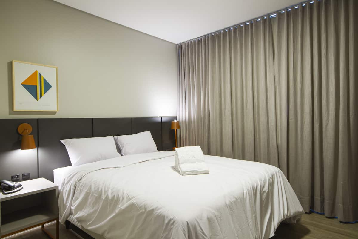 Habitación de hotel con una cama bien hecha, una lámpara de noche, una obra de arte enmarcada y un amplio ventanal con cortinas.
