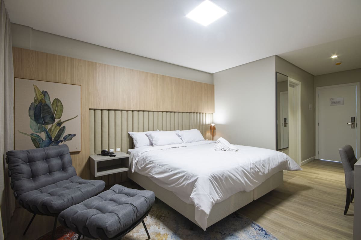 Una habitación de hotel moderna con una cama grande, un sillón gris, una mesita de noche y un cuadro en la pared.