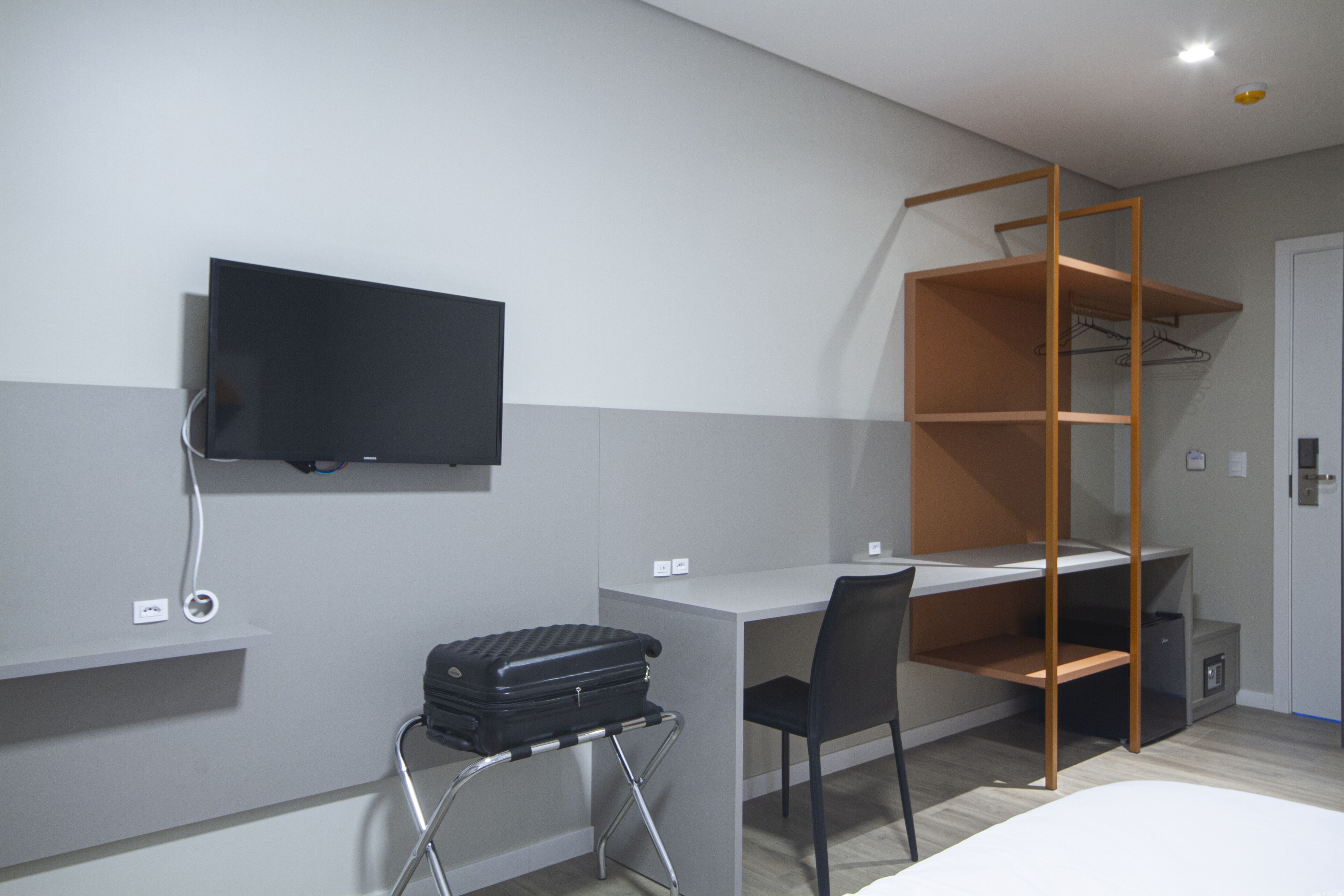 A modern hotel room with a desk, a television mounted on the wall, a suitcase on a chair, and a wardrobe with shelves.