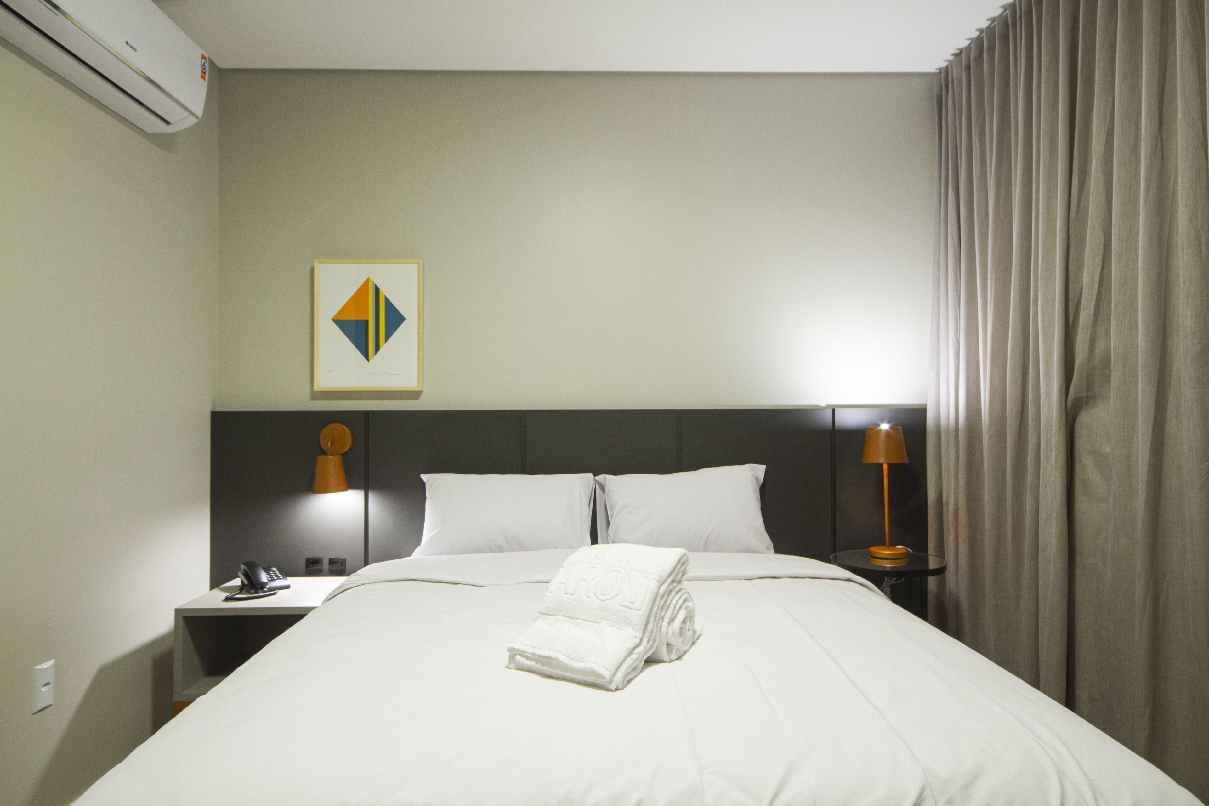 A hotel room with a neatly made bed, a nightstand with a lamp, a phone, and a wall-mounted air conditioner.