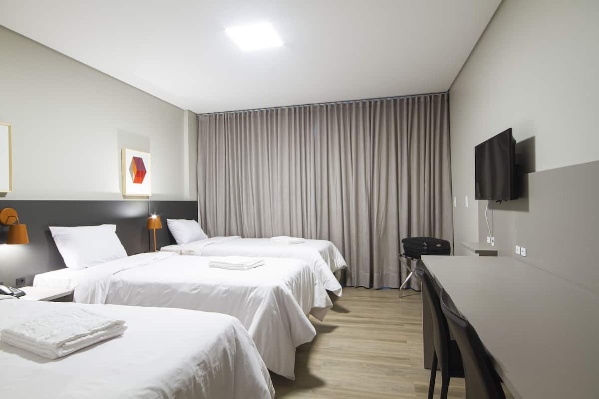 Una habitación de hotel con tres camas, un escritorio, un televisor y un amplio ventanal con cortinas.